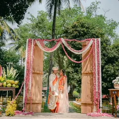 The couple stands at the entrance of their outdoor ceremony space. The wooden arch is draped with delicate pink and white floral garlands, setting a romantic tone for the vows.