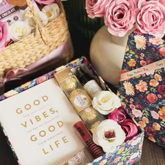 A thoughtful Mother's Day gift featuring the book "Good Vibes, Good Life," a Kama hydrating cream, Ferrero Rocher, a Nykaa lip color, and a Bath & Body Works hand cream, all in a floral printed box.