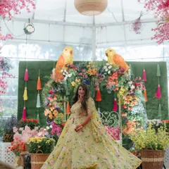 A bride twirls in her vibrant lehenga against a backdrop of colorful florals and parrot motifs, perfectly capturing the joyful spirit of a Mehendi function.