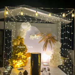 A versatile cabana-style backdrop illuminated with fairy lights and Edison bulbs. Decorated with gold and white balloon garlands and boho elements like dried palm fronds, this setup is suitable for birthdays, anniversaries, or even a bachelorette party.