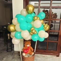 A unique "hot air balloon" decoration for a welcome baby party. The basket holds a cute teddy bear, and the balloon is made from a mix of teal, white, and gold balloons.