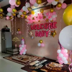 A festive "Welcome Baby" room decoration for a baby girl. The setup includes pink and gold foil letter balloons, ceiling balloons with ribbons, and balloon pillars.