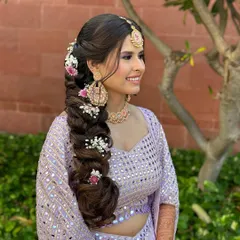 Mehendi ready! This intricate, voluminous braid is decorated with delicate pink and white flowers, perfectly complementing the bride's lavender lehenga for a soft, romantic look.