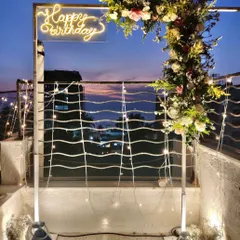 For this terrace birthday party, I created a modern and chic atmosphere with a custom neon "Happy Birthday" sign on a floral arch, set against a beautiful twilight city view. The setup includes fairy lights and lanterns for a warm, inviting glow.