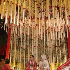 A floral haven for a sacred union. This mandap features an ethereal cascade of red roses and delicate white floral strings, with chandeliers casting a warm glow as the couple partakes in the wedding rituals.
