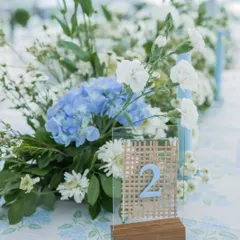 A table number with a woven cane detail, set against a floral arrangement of blue hydrangeas and white carnations, reflecting a modern-rustic aesthetic.