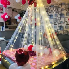 A cozy and romantic corner setup. This decoration features a fairy light canopy over the bed, with red and white balloons, including heart-shaped foils, creating an intimate and warm atmosphere for a special night.