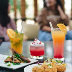 Here, the focus is on a table laden with colorful food and drinks, with guests blurred softly in the background. This composition creates a lively, social atmosphere, showcasing the menu in a real-world setting.