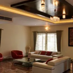 A modern living room featuring a striking black high-gloss false ceiling with integrated cove lighting and a chandelier. The space is furnished with minimalist white and red leather sofas, a glass coffee table, and set on a rich, red-toned marble floor.