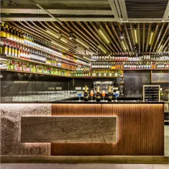 The exquisite bar counter at Pasta Street, crafted with a mix of stone veneer and fluted wood, is highlighted by dramatic backlighting. Above, a ceiling of varied lights and a fully stocked display create a mesmerizing focal point for the lounge area.
