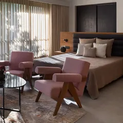 In this bedroom, natural light floods the space, highlighting the soft pink tones of the armchairs and the rich texture of the wooden headboard. The design creates a warm, inviting, and peaceful atmosphere.