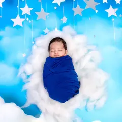 A dreamy newborn portrait with the baby swaddled in blue, floating on a soft cloud under a sky of stars. This is one of our most popular creative newborn photo ideas.
