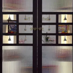 A stunning pooja room door made of dark wood and fluted glass, with small square openings adorned with traditional brass bells.