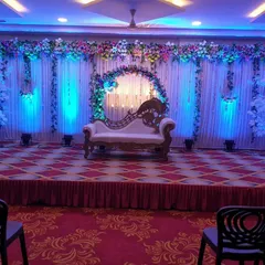 A classic engagement stage setup in a banquet hall. The decor includes a floral arch, a white backdrop with blue uplighting, and a comfortable diwan for the couple, creating a simple yet beautiful setting.
