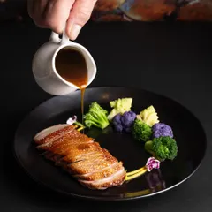 A fine-dining plate of sliced duck breast with a sauce being poured, capturing a moment of culinary action and elegance.