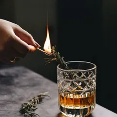 An action shot of lighting a sprig of rosemary for a smoked cocktail. The flame and the dramatic lighting create a sense of theatre and sophistication.