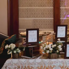 An elegant buffet setup for a birthday celebration, featuring mirrored stands, white floral arrangements, and framed menu cards for a clean and sophisticated look.