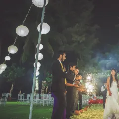The bride's entrance at an outdoor evening wedding, with guests holding sparklers to light her way. The paper lanterns and palm trees create a magical, romantic atmosphere.