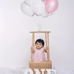 A dreamy and classic first birthday studio portrait. The hot-air balloon basket, cloud-like floor, and soft pink balloons create a whimsical atmosphere perfect for celebrating your baby's special day.