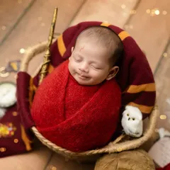 A tiny wizard in the making. This smiling newborn is wrapped snugly in a red blanket for our Harry Potter themed session, complete with a magic wand and a little owl friend.