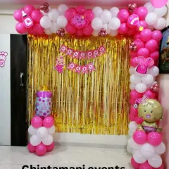 A sweet and simple 'Welcome Baby Girl' decoration for a home. The pink and white balloon arch frames a gold tinsel backdrop, and I've added baby-themed props like a bottle and rattle to complete the look.