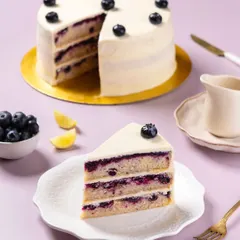 A slice of a multi-layered blueberry and lemon cake, photographed for The Big Chill Cafe. The focus is on revealing the cake's internal structure, showing the fresh fruit filling and soft sponge, a key shot for any bakery or dessert menu.