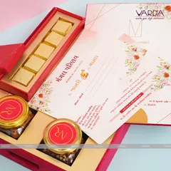 This luxurious red velvet box is filled with gourmet delights, including jars of flavored nuts and a separate compartment for gold-wrapped sweets. The accompanying invitation card features a beautiful floral border.