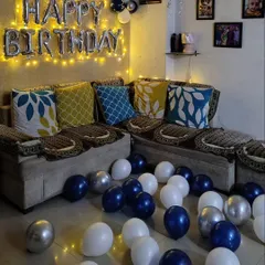 A simple and elegant birthday setup in a living room corner. This decoration uses blue, white, and silver balloons on the floor and a 'Happy Birthday' foil sign lit with fairy lights for a cozy celebration.