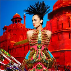 A powerful fashion image that blends Indian motifs with avant-garde design. The model stands before a vibrant red backdrop of the Red Fort, wearing a fantastical, structured garment.