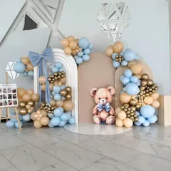 A sweet and classic teddy bear theme for a first birthday. The backdrop features arched panels, a large teddy bear cutout, and a beautiful balloon garland in pastel blue, beige, and gold.