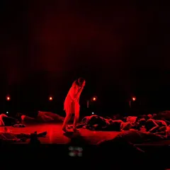 Bathed in red light, a solo performer stands amidst a group in a scene depicting struggle and resilience. This highlights our use of lighting and stage composition to evoke strong emotions and tell a compelling story through dance.