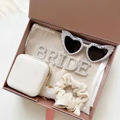 A celebratory gift box for the soon-to-be bride. This hamper includes a 'BRIDE' pouch with pearl lettering, fun heart-shaped sunglasses, a jewelry case, and a scrunchie, perfect for her bridal shower.