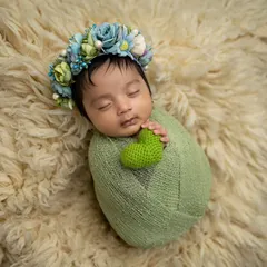 A classic 'potato sack' pose. The baby is snugly wrapped in a green swaddle, holding a tiny heart, and resting on a soft fur rug. This pose is great for keeping babies calm and sleepy.