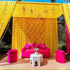 This is a bright and sunny haldi decoration with a canopy of yellow drapes. The backdrop features a crisscross pattern of marigold strings, and the vibrant pink sofa adds a lovely contrast for a fun photo corner.