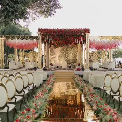 A magnificent mandap setup at The Leela Palace Udaipur, with a mirrored aisle reflecting the sky. The design features a rich palette of red florals and elegant white seating, creating a regal atmosphere for the lakeside ceremony.