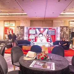 The main event space for a Bollywood-themed 90th birthday party. The setup includes a custom stage designed to look like a film set, cocktail tables with sequined cloths, and a large screen for presentations.