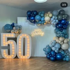 A full view of the decor for a 50th birthday party. The combination of large marquee numbers, a neon sign, and a sophisticated blue and beige organic balloon garland creates an impressive focal point for the event.
