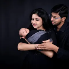 A beautiful composition of a new family. The mother holds their sleeping newborn while the father stands behind, looking over them with a gentle smile, all set against a classic dark backdrop.