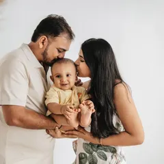 A loving family portrait with both parents kissing their happy baby's head. The neutral tones and simple props create a warm and timeless feel.