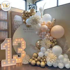 A chic and modern setup for a 13th birthday. The decor features a neutral palette with white and gold balloons, pampas grass, and large light-up marquee numbers.
