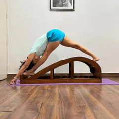 This is a supported Paschimottanasana (Seated Forward Bend) using a backbend bench. The gentle slope of the bench helps to lengthen the spine and release the hamstrings without force, making it a deeply restorative forward extension.