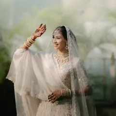 A happy bride in her all-white wedding lehenga, waving with a smile. The soft, diffused light gives the portrait an ethereal quality.
