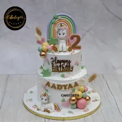 An elegant two-tier unicorn cake in a soft white and pastel theme. This eggless blueberry cake features two adorable fondant unicorns, a colorful rainbow, and decorative balloons, perfect for a second birthday.