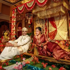 A royal portrait of the couple, seated amidst the opulent decor of their South Indian wedding. The golden mirror floor reflects the rich floral arrangements, creating a truly grand and elegant setting for their special day.