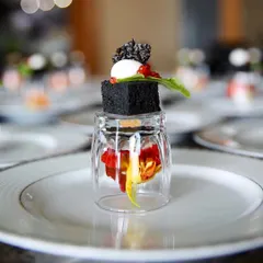 A deconstructed appetizer served in a unique way, with a savory black cube balanced on an inverted glass containing edible flowers. This is an example of our innovative and multi-layered plating style.