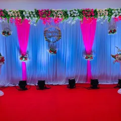 An elegant and simple stage decoration with a white curtain backdrop and tasteful pink drapes. A floral arrangement hangs from the top, and a custom wooden hoop with the couple's names adds a personal touch to this reception setup.