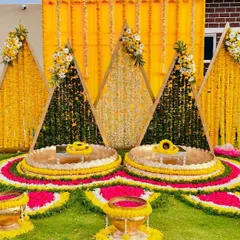 A truly unique Haldi seating arrangement. This setup features triangular backdrops made of marigold flowers and lush greenery, with intricate floral rangoli on the ground for a stunning visual effect.