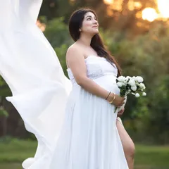 An outdoor maternity portrait taken during the golden hour, with soft sunlight creating a warm, ethereal glow. The flowing white gown and natural setting provide a romantic and serene feel.