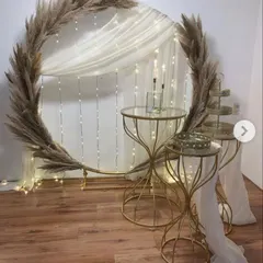 This is a modern and minimalist backdrop perfect for a cake-cutting ceremony or a photo corner. It features a large golden ring decorated with pampas grass, with a backdrop of sheer curtains and fairy lights for a soft, dreamy effect.