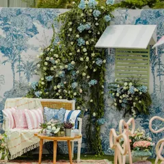 An aesthetic photo booth from a 'Carnival of Love' theme. The blue and white toile backdrop, combined with lush florals and elegant seating, creates a picture-perfect, European-inspired corner.